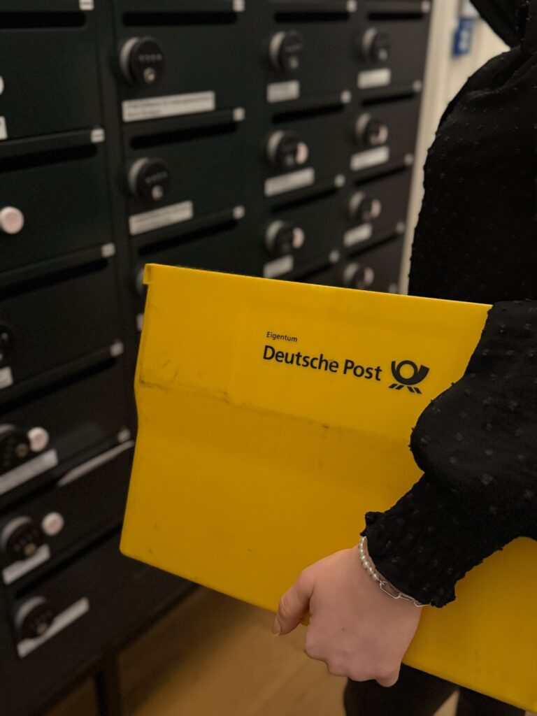 Eine Person hält eine gelbe Deutsche Post Kiste vor gesicherten Postfächern.“